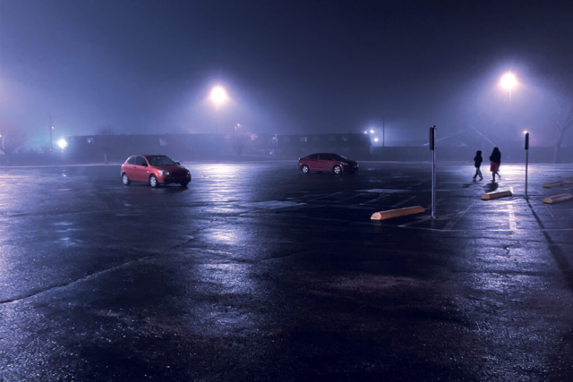 Photo of a parking lot in the dark. Two red cars are parked across the lot from each other and two people can be seen walking in the direction of the cars